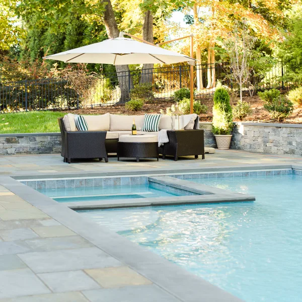 outdoor lounge area features wicker seats and off-white cushions under a large umbrella near an underground pool